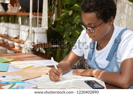 Sideways shot of dark skinned lady in casual clothes, writes down in notebook, checks paper graphics, works freelance, wears optical glasses, edits text on free time. People and work concept