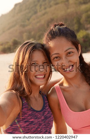 Smiling mixed race female friends pose near for making selfie, dressed in top, have leisure time for sport, model against cliff background, enjoy summer holidays. Friendship, relationships concept