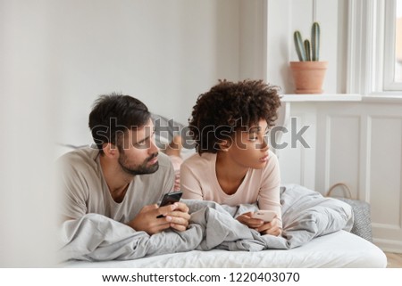 Contemplative family couple use cellulars, concentrated aside, lie in bed at morning, connected to wireless internet at home, text messages, invite friends for party, ignore live communication