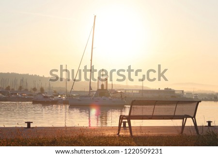 The sailing yacht enters under the motor in the marina in the rays of the morning sun. Dawn in the port. Empty bench on the pier against the lighthouse. Symbols of return and waiting.