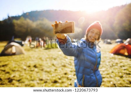 happy smiling woman wearing in a blue down jacket and a red cap is standing in campsite on a meadow in the autumn forest on a bright sunny foggy morning and taking selfie photo on the phone camera.