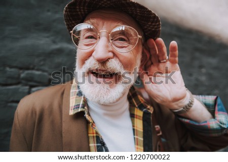 Concept of positive mood lifestyle of hearing impaired person. Close up portrait of happy pensioner with playful glance holding palm near his ear Royalty-Free Stock Photo #1220700022