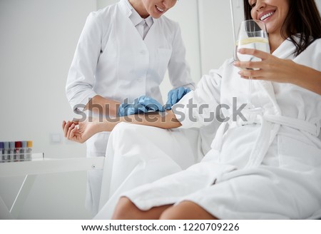 Cropped portrait of charming woman in white bathrobe sitting in armchair and receiving IV infusion. She is holding glass of lemon beverage and smiling Royalty-Free Stock Photo #1220709226