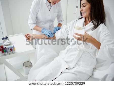 Cropped portrait of beautiful woman in white bathrobe sitting in armchair and receiving IV infusion. She is holding glass of beverage with lemon and smiling Royalty-Free Stock Photo #1220709229