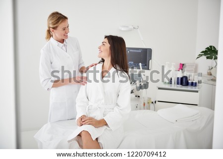 Portrait of charming woman in white bathrobe sitting on daybed and looking at beautician with smile while she touching her shoulder Royalty-Free Stock Photo #1220709412
