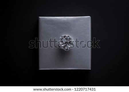 Square shape gift wrapped in silver paper, decorated with silver pine cone on black background