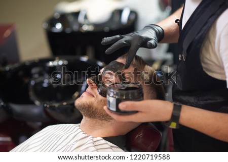 Side view of smiling young man lying on chair in vintage barber shop while barber in protective gloves putting black mask against dots on face. Concept of skin care and cleaning. Royalty-Free Stock Photo #1220719588