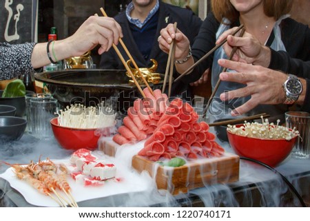 Asian Hot pot surrounded by delicios food in dry ice, people eating with chopsticks. Translation of hot pot and wall inscription: "hogo hot pot", "
singing is heard under the roof of the house"