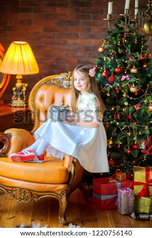 beautiful little girl in a dress sits in an armchair with a gift in hand, near the holiday tree