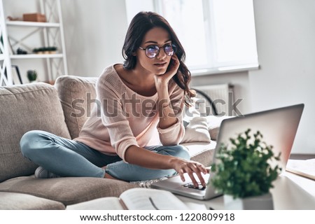 Surfing the net. Thoughtful young woman in eyewear using computer while sitting on the sofa at home                  Royalty-Free Stock Photo #1220809918