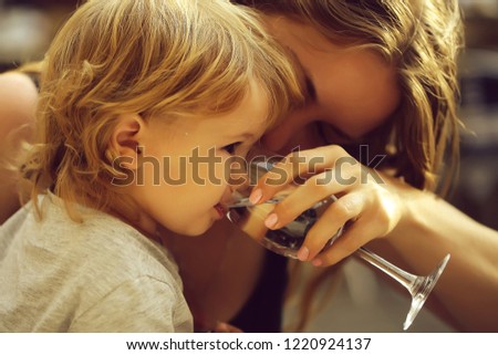 Smiling young mother holding glass and child boy with blonde hair drinking water from paret hands sunny day outdoor, horizontal picture