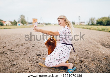 Nice looking beautiful blonde girl making selfie photo on smartphone. Young happy pretty female in stylish retro dress riding toy horse outdoor. Strange bizarre fashionable woman portrait.  Childhood
