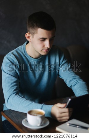 Guy is sitting in a cafe and using a tablet. Student spend time in cafe and drink coffee. He study and note helpful information