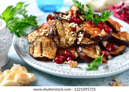 Grilled eggplants in tomato sauce with walnuts and pomegranate seeds on a blue plate on a light slate, stone or concrete background.