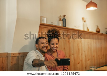 Smiling young couple sitting at a table in a trendy bar laughing and taking a selfie together 