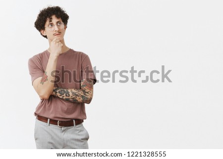 To be or not that is question. Thoughtful artistic and creative smart curly-haired guy in glasses with moustache and cool tattoos on arms rubbing chin gazing up while thinking picturing plan in mind