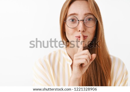 Close-up shot of tender and feminine cute redhead female in glasses with freckles showing shh sign making shush sound with index finger over mouth smiling mysteriously with love and care