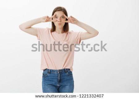 Studio shot of emotive carefree young female friend in pink t-shirt with tattoos on arms, showing disco sign over eyes, pouting, making childish expression while having fun and enjoying great day