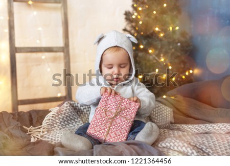 A little boy is sitting with a gift on the bed on the background of the Christmas tree and the stairs with a garland. New Year, Christmas card in a Scandinavian interior. Picture with highlights 