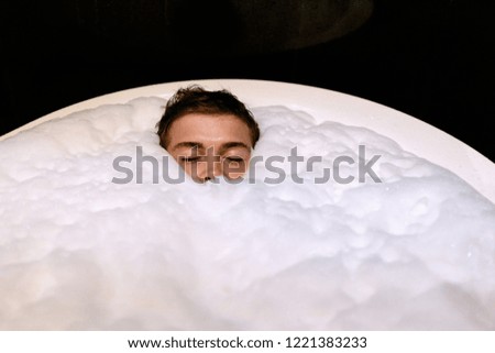 Person is relaxing in a round bath full of white foam. Minimalistic photo about spa with free space for your text. Man with eyes closed deep in bath foam