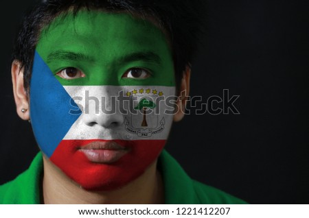 Portrait of a man with the flag of the Equatorial Guinea painted on his face on black background, tricolor of green white and red with a blue triangle and the Coat of arms.