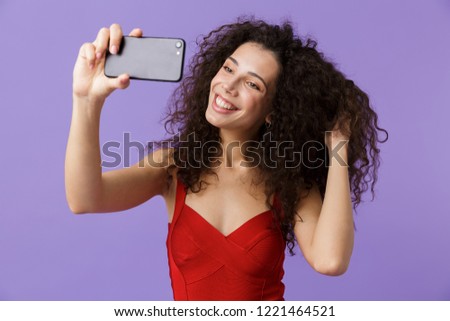 Image of curly woman 20s wearing red dress taking selfie photo on black smartphone standing isolated over violet background