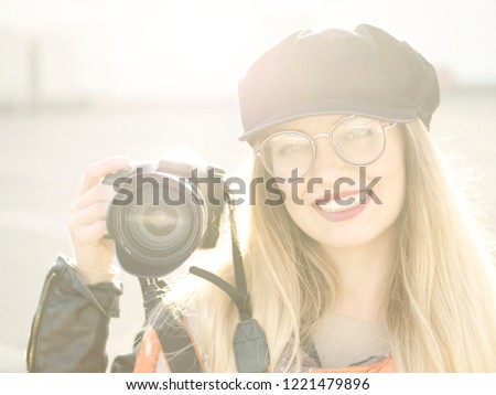 Portrait of a beautiful stylish cheerful young blonde photographer in glasses and a hat with makeup holding a black camera in a cookie on a light background
