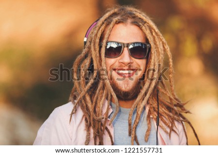 Portrait of hipster blonde bearded boy with dreads wearing headphones and glasses walking and smiling outdoors an the sunny autumn day