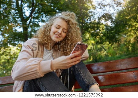Young beautiful girl blonde with curly hair sits with her legs on a park bench, listens to her favorite music in headphones with eyes closed from pleasure, wearing a pink jacket and black pans