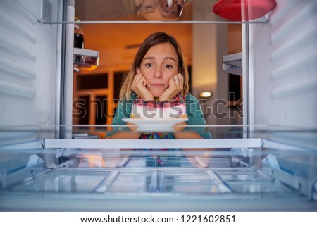 Woman standing in front of fridge with head in hands and looking at cheesecake. Picture taken from the inside of fridge.