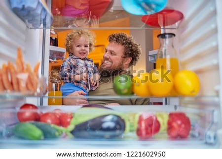 Father and son standing in front of opened fridge and looking something to eat at night. Fridge full of groceries. Picture taken from the inside of fridge.