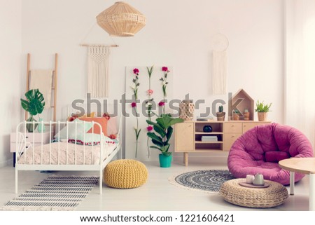 Stylish pink settee in the middle of elegant boho bedroom with macrame on the wall, flower board and single metal bed with pillows, real photo