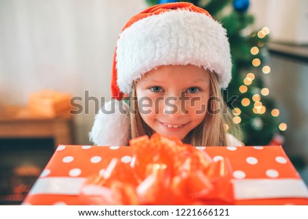 Picture of small and beautiful girl holding read box with present and looks on camera. She is happy. Girl wears Christmas hat on head. There is tree behind her