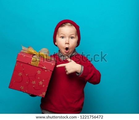 Funny little hipster in red Santa hat. Child with Christmas gift. Stylish boy kid against blue wall at home. Happy xmas and New Year, winter holiday concept