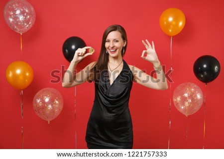 Pretty woman in black dress showing OK sign, holding bitcoin, metal coin of golden color, future currency on bright red background air balloons. Happy New Year, birthday mockup holiday party concept