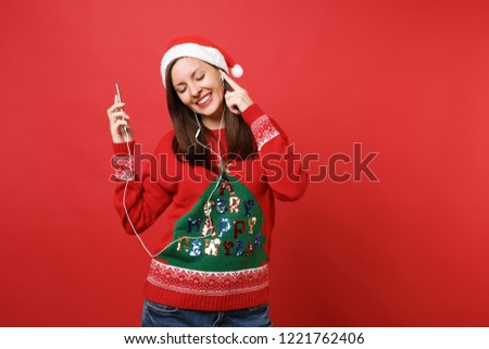 Calmed young Santa girl with earphones keeping eyes closed, holding mobile phone, listening music isolated on red background. Happy New Year 2019 celebration holiday party concept. Mock up copy space