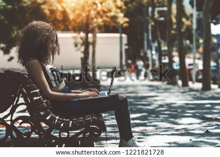 A concentrated charming female freelancer is sitting on a wooden bench in a park, using the netbook; beautiful undergraduate girl in glasses and with bulky curly hair is working on her laptop outdoors