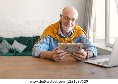 Indoor image of sentimental bearded bald grandfather wearing spectacles and elegant clothes sitting at wooden desk in front of open laptop, holding photo frame, looking at picture of his children