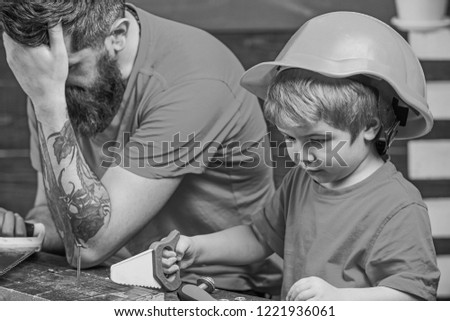 Educational games concept. Boy, child busy in protective helmet learning to use handsaw with dad. Father, parent with beard looks dissapointed while son play with toy saw.