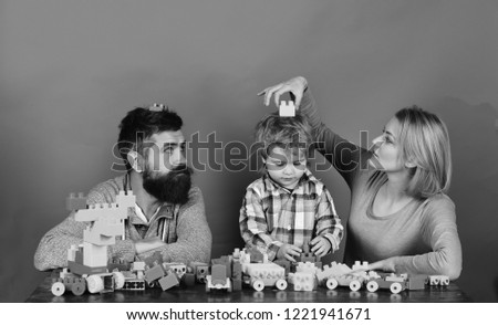 Man with beard, woman and boy play on red background. Family with serious faces build toy cars out of colored construction blocks. Mom, dad and kid in playroom. Childhood and playing concept.