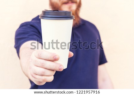 Young man holds disposable white paper cup of coffee. Copy space on cup.