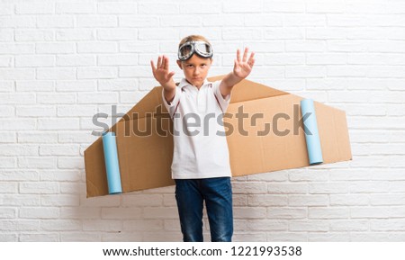 Boy playing with cardboard airplane wings on his back making stop gesture with her hand