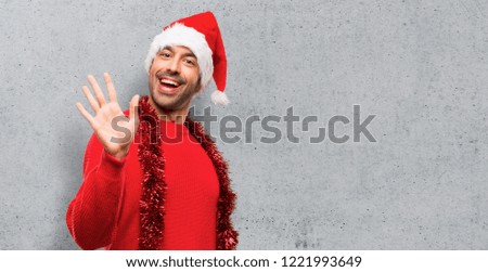 Man with red clothes celebrating the Christmas holidays saluting with hand with happy expression on textured background