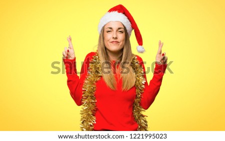 Blonde woman dressed up for christmas holidays with fingers crossing and wishing the best on yellow background