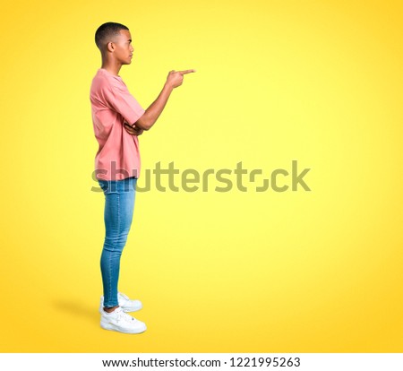 Standing young african american man pointing finger to the side and presenting a product in lateral position on colorful background