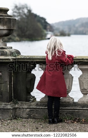 A young girl in a red coat stands staring overlooking a beautiful large lake leaning on a stone wall