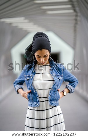 Portrait of a tall, young, elegant and attractive Malay Muslim woman standing alone on a beautiful bridge during the day. She is wearing a white dress, denim jacket and head scarf. 