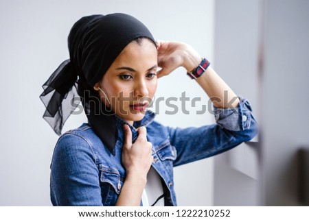 Portrait of a young, attractive and fashionable Middle Eastern Muslim woman in a hijab head scarf. She is standing by the window and is enjoying the sun as she takes a break. 