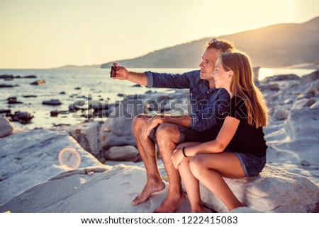 Father and daughter relaxing on a rocky beach by the sea and taking selfie with smart phone