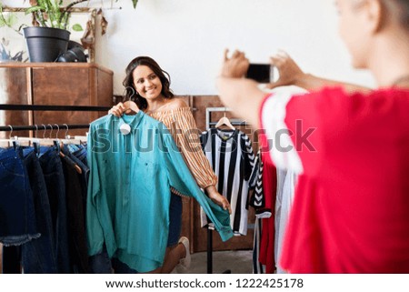 friendship, shopping and technology concept - woman taking picture of happy friend by smartphone at vintage clothing store